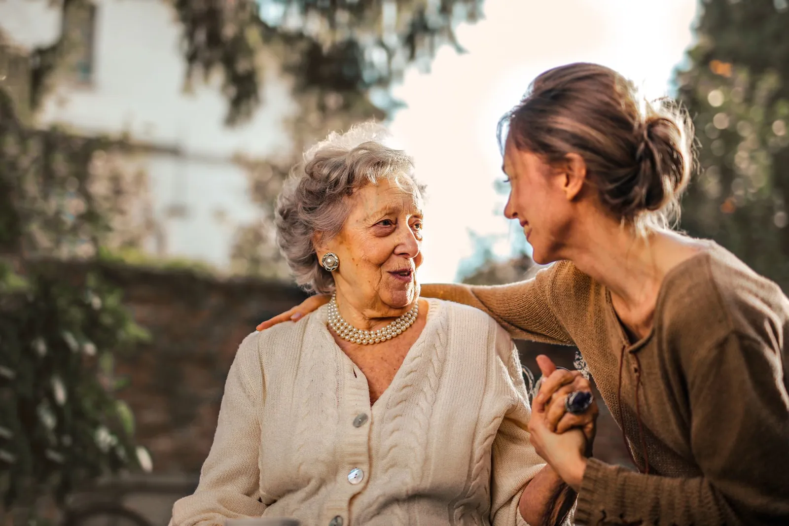 A support worker and an older woman clasp hands, laughing together in a garden