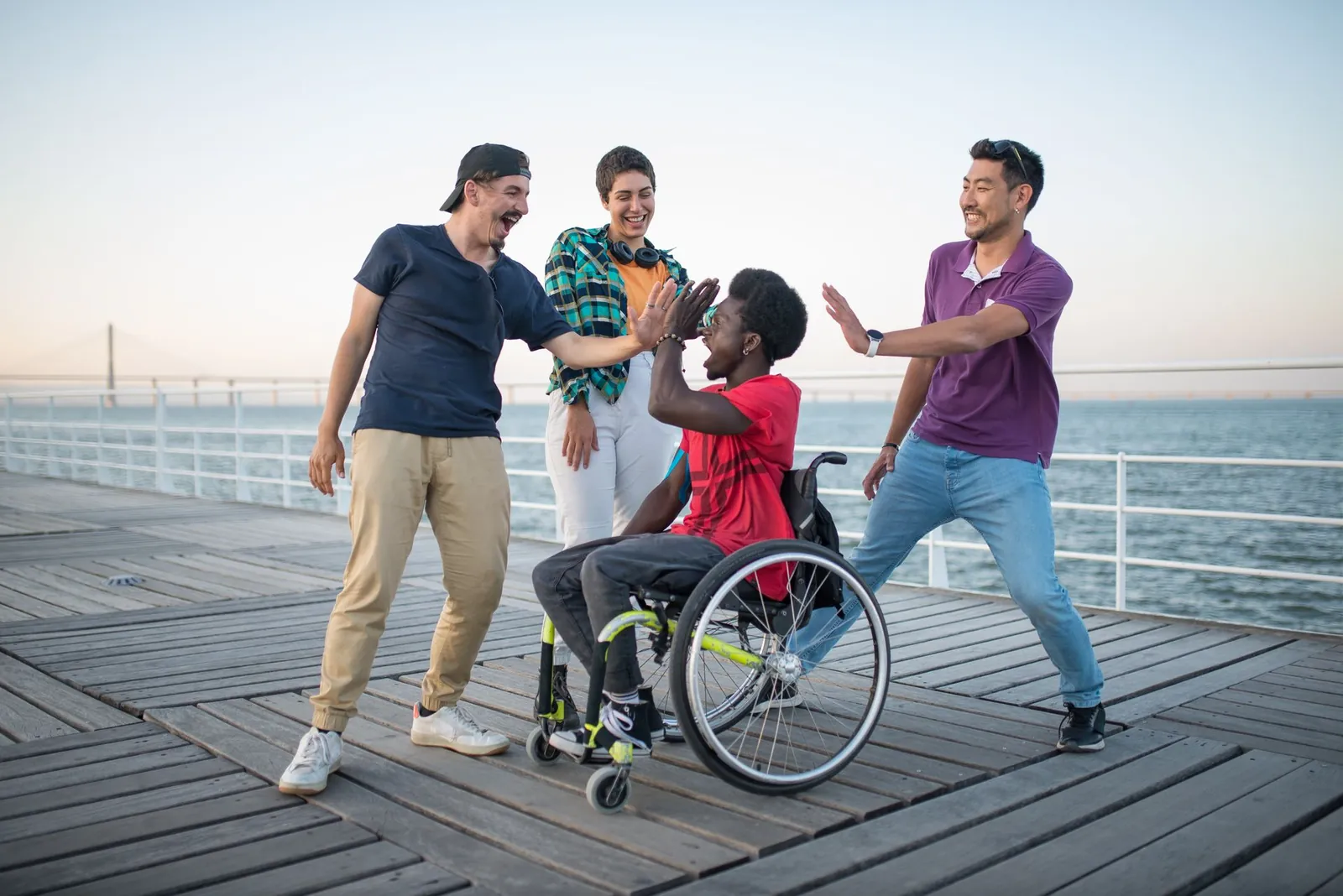 Friends celebrate with a high-five on a seaside pier, one of them using a wheelchair