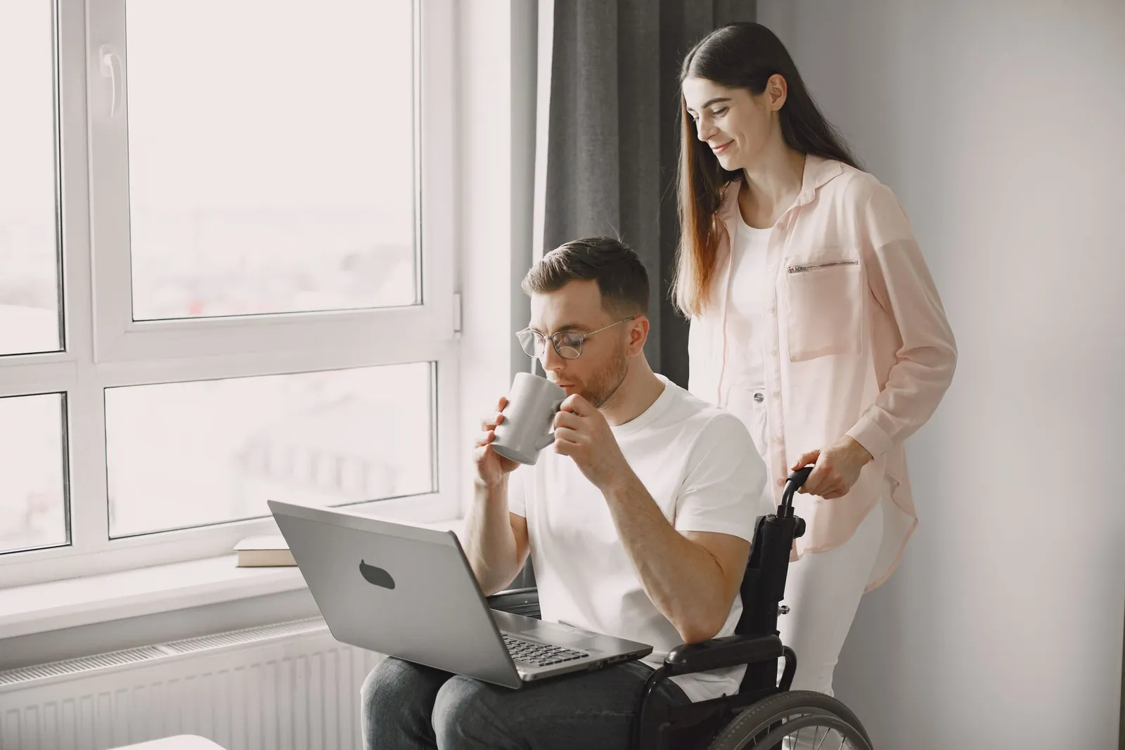 A support worker and a man using a wheelchair share coffee and a laugh over a laptop at home